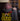 A person sits at a table, head down and arm covering their face, appearing overwhelmed. The Hot Ones logo with a flaming chicken is overlaid on the image.