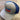 The Blue/Grey Puckerbutt Snapback Hat, featuring an embroidered PuckerButt Pepper Company logo with a chili pepper design, rests on a wood-textured surface.